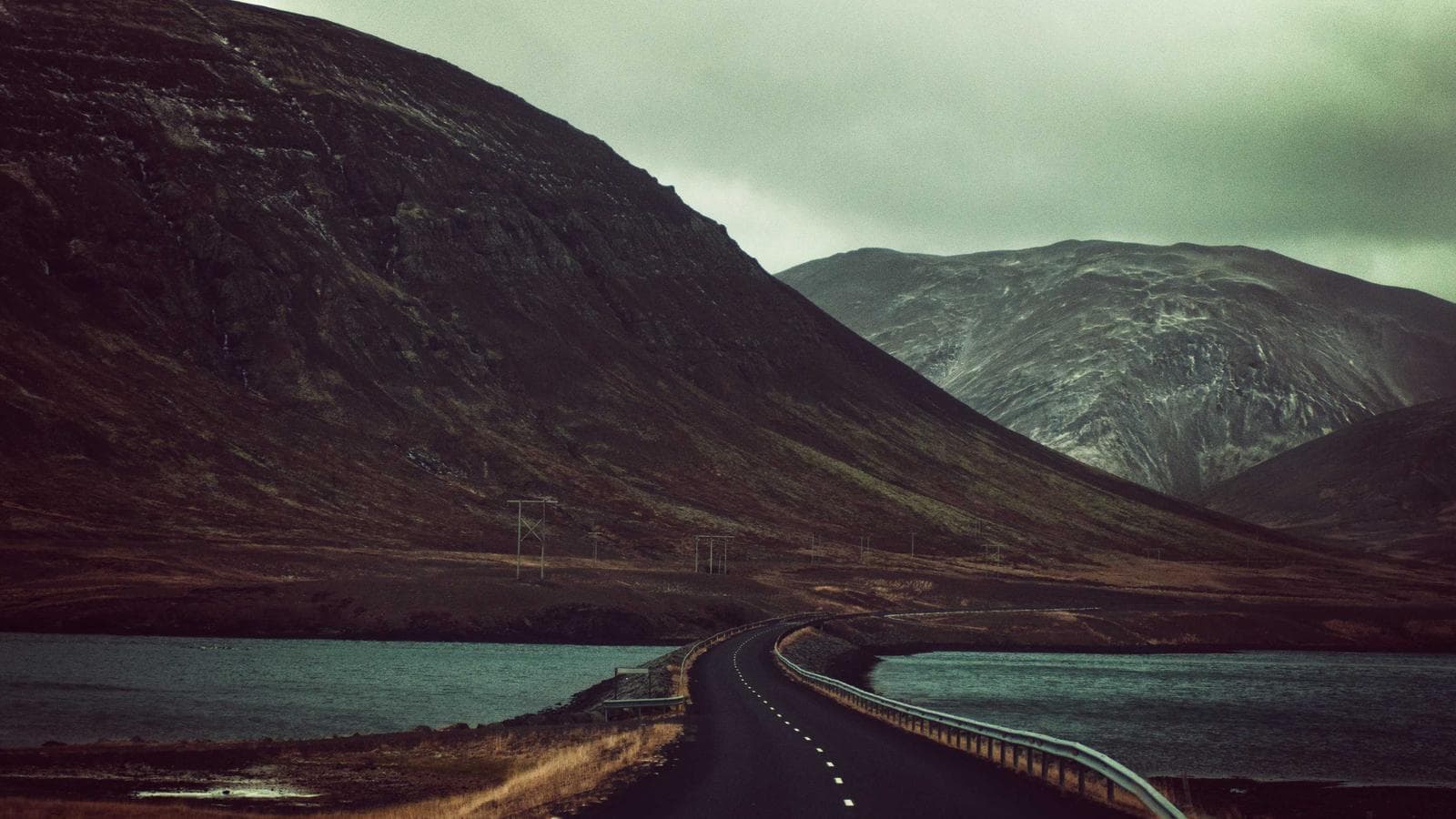 A desolate road curves over a body of water, flanked by dark, imposing mountains under a cloudy sky.