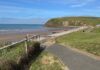 Places to Go: St Bees A tarmac path running down grassy ground towards the sandy beach at St Bees, with the green headland of St Bees Head rising beyond and the Irish Sea to the left