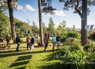 Communities in Focus Exhibition Opens at Willow Burn Hospice Gardens Visitors walking through Willow Burn Hospice Gardens in evening sunshine, with framed portraits from the Communities in Focus exhibition hung on lines strung between the trees