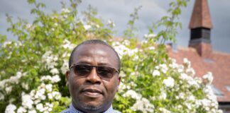 A Century of Faith and Community at All Saints Church Rev. Fr. Vernatius Edih, Parish Priest of All Saints RC Church, standing outside the church in a clerical shirt and collar in front of a white rose bush