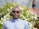 A Century of Faith and Community at All Saints Church Rev. Fr. Vernatius Edih, Parish Priest of All Saints RC Church, standing outside the church in a clerical shirt and collar in front of a white rose bush